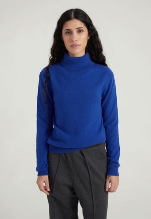 Blue turtleneck sweater made of soft material, features long sleeves and a relaxed fit, paired with dark gray pants.