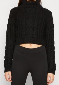 Schwarzer, kabelgestrickter, kurz geschnittene Pullover mit hohem Kragen, der strukturierte Ärmel und einen gerippten Saum hat, kombiniert mit schwarzen, gerippten Leggings.
