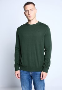 Pull en tricot vert avec col rond, poignets et ourlet côtelés. Présente un petit détail de marque sur la poitrine. Associé à un pantalon en denim bleu.