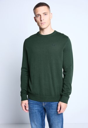 Suéter verde de punto con cuello redondo, puños y dobladillo acanalados. Presenta un pequeño detalle de marca en el pecho. Combina con pantalones de mezclilla azul.
