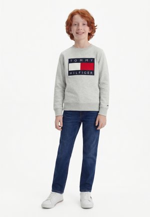 Dijete s crvenom kosom stoji, nosi sivu Tommy Hilfiger majicu, plave traperice i bijele tenisice, smiješi se u kameru.