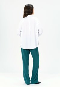 Camisa blanca de manga larga con cuello alto, combinada con pantalones anchos de pierna ancha en color teal, que muestran una textura suave y un diseño minimalista.