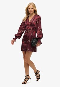 Vestido floral burdeos con un escote en V profundo, mangas largas abullonadas y cintura ajustada. Combinado con tacones negros con tiras y un clutch negro texturizado.