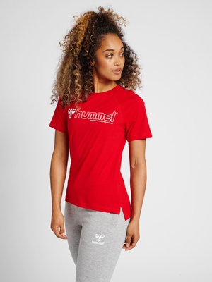 Kobieta z kręconymi włosami, ubrana w czerwoną koszulkę Hummel i jasnoszare spodnie dresowe Hummel, patrzy w lewo na jednolitym tle.