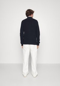 Námornícky pletený sveter s vysokým límcem a žebrovanou texturou, spárovaný s volnými bílými kalhotami. Oblečení je jednoduché a neformální, viděné zezadu.