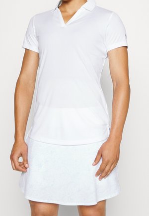 Persona con una camisa polo blanca de manga corta y una falda blanca con sutiles patrones florales, de pie con los brazos relajados.