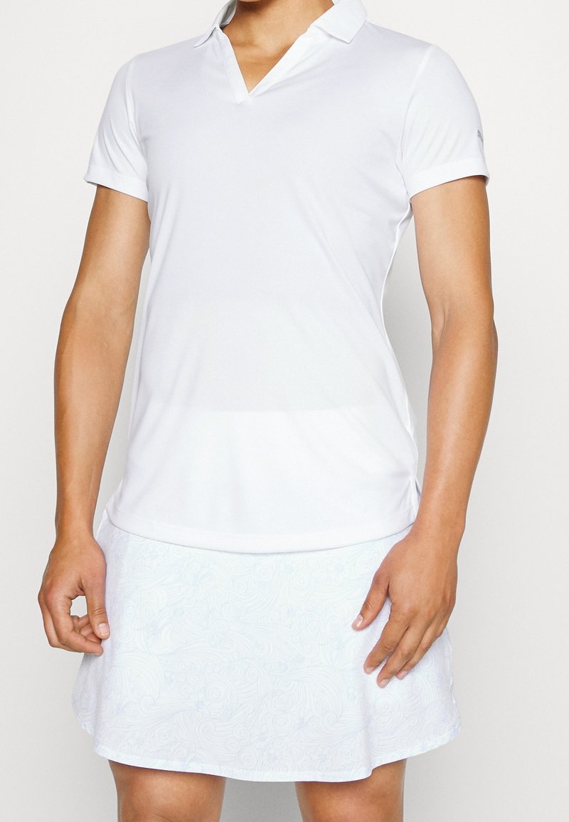 Personne portant un polo blanc à manches courtes et une jupe blanche avec des motifs floraux subtils, se tenant les bras détendus.