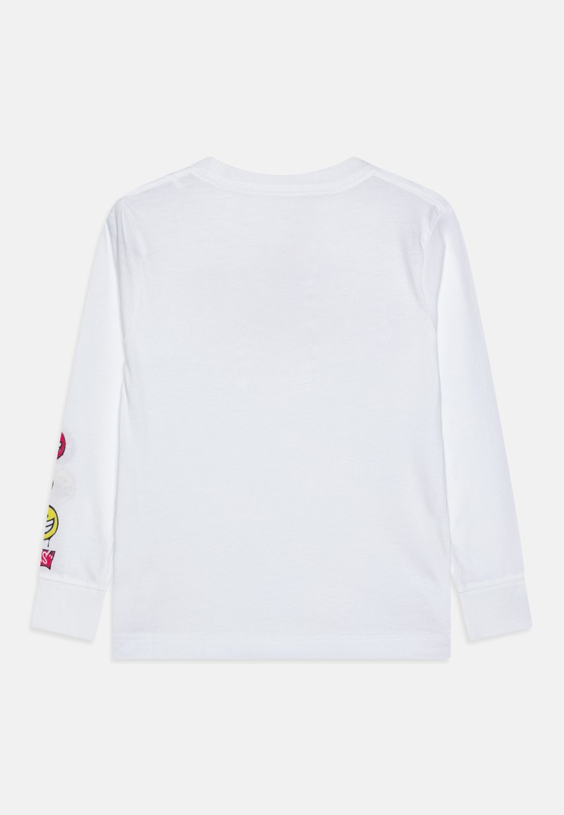Camisa blanca de manga larga colocada plana, con el lado de atrás visible, y parches de caras sonrientes coloridas en la manga izquierda cerca del puño.