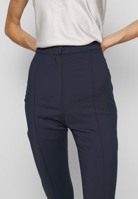 Marineblauwe getailleerde broek van gladde stof met een middelhoge tailleband, voorzien van een naaddetail aan de voorkant en zonder zichtbare zakken.