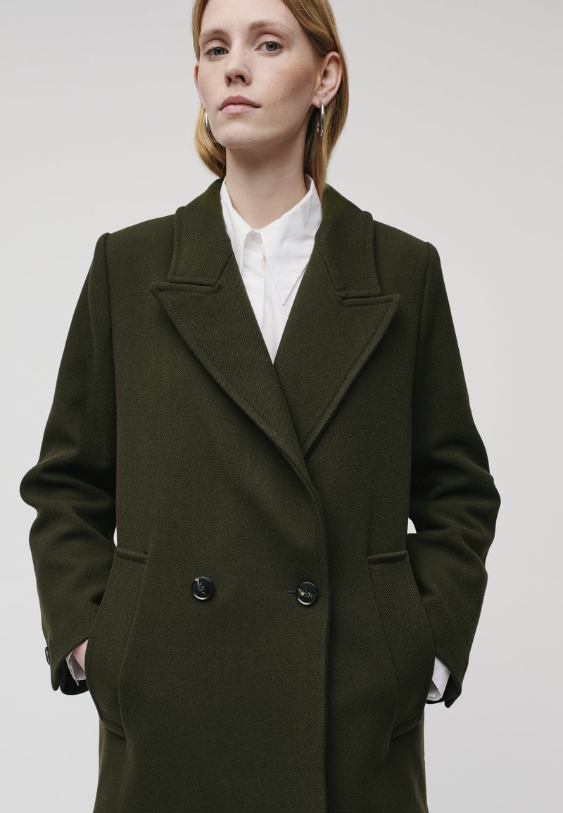 Manteau olive vert croisé avec revers, texture douce, grandes poches avant et boutons noirs sur une chemise blanche à col.