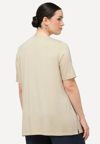 Beige Kurzarmoberteil mit lockerer Passform, glatter Textur, abgerundetem Saum und seitlichen Schlitzen. Die Rückansicht zeigt schlichte Nähte und einen runden Ausschnitt.