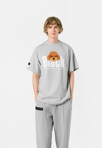 Camiseta ligera de gran tamaño en gris claro, con un gráfico de un perro marrón texturizado y texto en blanco audaz. Combinada con unos pantalones grises claros a juego.