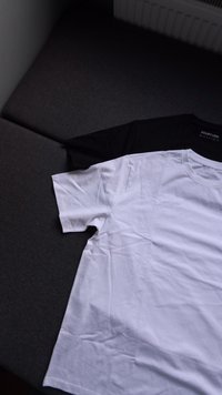 Due t-shirt in cotone distese su una superficie piatta: una bianca e una nera, con maniche corte e collo rotondo. Texture liscia, design minimalista, senza motivi.