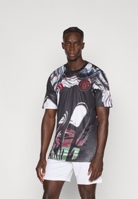 Camicia nera a maniche corte con motivi astratti multicolori e un logo circolare rosso sul petto, abbinata a pantaloncini bianchi.