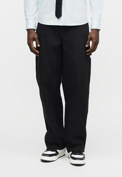 Άτομο που φοράει μαύρο παντελόνι με φαρδιά πόδια, λευκά αθλητικά παπούτσια με μαύρες λεπτομέρειες, λευκό πουκάμισο με κουμπιά και μαύρη γραβάτα, σε απλό λευκό φόντο.