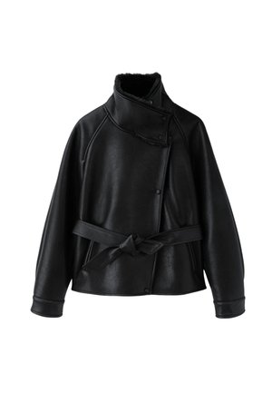 Femme portant une veste en cuir noire avec col en shearling, un débardeur blanc, un pantalon ample beige avec une ceinture noire et des chaussures noires, posant sur fond gris.