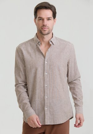 Mand iført en beige og hvid vertikalt striped langærmet skjorte og brune bukser, stående mod en ensfarvet lys baggrund.