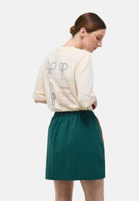 T-shirt crème à manches courtes avec des motifs graphiques au dos, associé à une jupe verte à taille élastique avec une poche latérale et un accent orange.