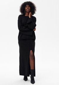 Kobieta o kręconych włosach, ubrana w czarny sweter z długim rękawem, czarną maxi spódnicę z rozcięciem na udzie oraz czarne botki, stojąca na białym tle.