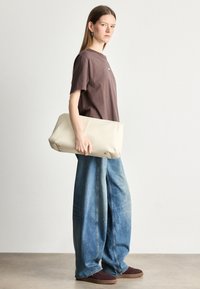 Beżowa torba w rozmiarze oversize o gładkiej fakturze i widocznych szwach, trzymana przez osobę w brązowym t-shircie i szerokich dżinsach, stojącą bokiem.