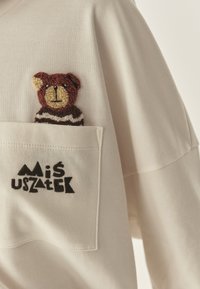 Jasnobeżowa koszula z długim rękawem, na której znajduje się haftowany brązowy miś o fakturze w kieszeni oraz czarny stylizowany napis "Miś Uszatek" poniżej.