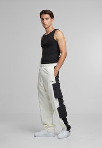 Black tank top, cream cargo pants with black side accents and multiple pockets, paired with white sneakers. Smooth fabric, relaxed fit.