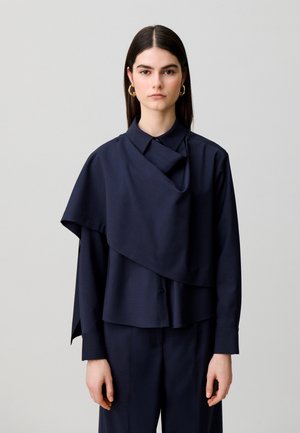 Jeune femme aux cheveux lisses foncés porte un chemisier bleu marine asymétrique à couches et un pantalon assorti sur un fond clair uni.