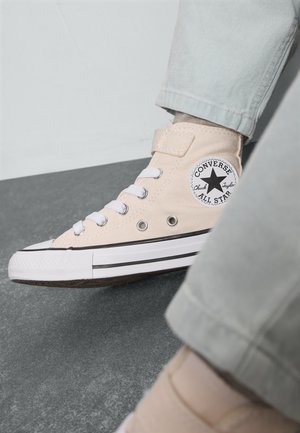Beige canvas hoge sneakers met witte veters, Chuck Taylor-patch, zwarte afwerking en rubberen zool, gedeeltelijk zichtbaar tegen een grijze achtergrond.
