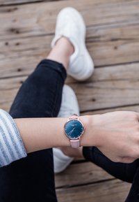 Okrągły zegarek z niebieską tarczą, różowozłotą kopertą i różowym skórzanym paskiem. Posiada minimalistyczne oznaczenia i małą koronkę.