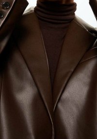 Zbliżenie na osobę w ciemnobrązowej skórzanej marynarce noszonej na dopasowanym brązowym swetrze z golfem, podkreślające fakturę i detale kołnierza.