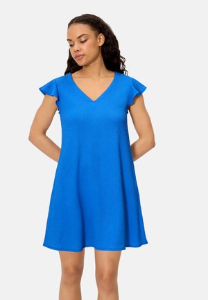 Mujer con cabello largo y rizado, vestida con un vestido azul de manga corta y cuello en V, de pie con las manos detrás de la espalda contra un fondo liso.