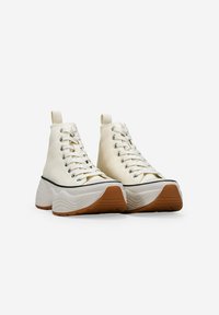 Zapatillas altas de lona en color off-white con una suela gruesa y ondulada y acentos de goma. Cuenta con ojales metálicos y cordones planos.