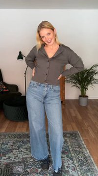 Jonge vrouw met licht haar die een grijze, dichtgeknoopte cardigan en wijde jeans draagt, staat glimlachend in een woonkamer met planten en meubels.