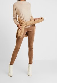 Suéter de punto beige combinado con pantalones marrones texturizados, atados con un envoltorio de punto. La modelo lleva botas blancas hasta el tobillo, que exhiben un diseño liso y ajustado.