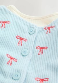 Ljusblå ribbad tyg med röd båge broderi. Har blå knappar längs framsidan. Mjuk textur och rund halsdetalj.