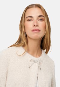 Kremowy, miękki kardigan z okrągłym dekoltem, o delikatnym połysku i ozdobnym, srebrnym zapięciem w kształcie kokardki z przodu.