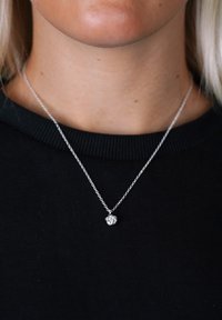Söt silverhalsband med en fin kedja och en liten, strukturerad berlock som liknar ett knutet rep, bärs över en svart T-shirt med rund hals.