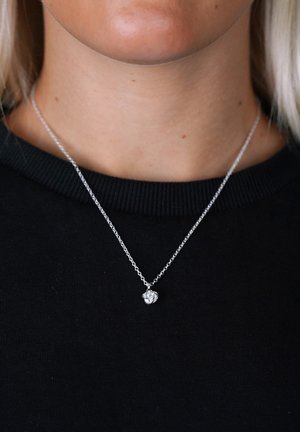 Söt silverhalsband med en fin kedja och en liten, strukturerad berlock som liknar ett knutet rep, bärs över en svart T-shirt med rund hals.