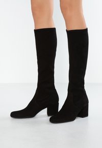 Bottes noires en daim montant jusqu'au genou, présentant un talon carré, des détails de coutures courbées et une silhouette ajustée le long de la jambe. Texture lisse.