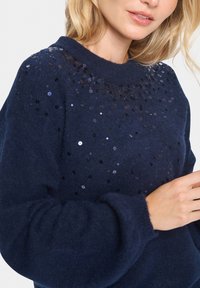 Pull marine en tissu doux avec un col côtelé. Présente des sequins noirs et argentés dispersés le long de la zone des épaules pour l'embellissement.