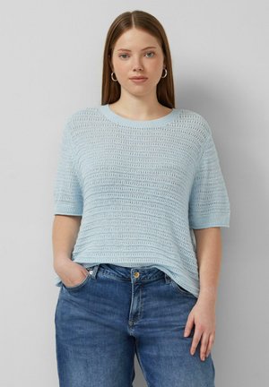 Młoda kobieta o prostych brązowych włosach, ubrana w jasnoniebieski dzianinowy top z krótkim rękawem i niebieskie dżinsy, stoi z jedną ręką w kieszeni na tle szarej ściany.