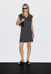 Grau gestricktes zweiteiliges Set mit ärmellosem Oberteil und V-Ausschnitt sowie passendem, figurbetontem Rock. Abgerundet mit schwarzen Schuhen und einer Handtasche.