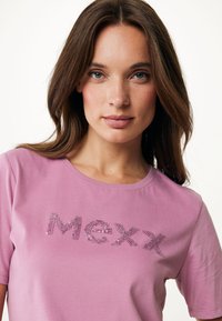 Růžové tričko s krátkými rukávy vyrobené z bavlny, které má na sobě flitrové logo "Mexx" z lesklých akcentů, kulatý výstřih a hladkou texturu.