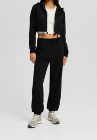 Svart kort hoodie med dragkedja och snörning, kombinerad med svarta sweatpants med resårmidja, och bärs med grå träningsskor.