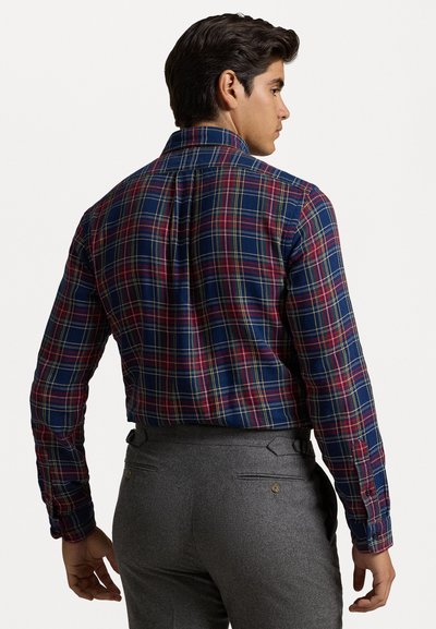 Chemise à carreaux dans les couleurs marine, rouge et jaune ; manches longues ; poignets boutonnés ; plis au dos ; associée à un pantalon gris.