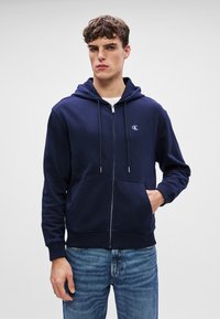 Hanorac bleumarin cu fermoar, cu glugă ajustabilă, fabricat din bumbac, cu buzunare frontale tip kangaroo și un logo mic pe piept. Purtați-l cu blugi albaștri.