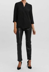 Blouse noire à manches trois-quarts avec un col en V et une finition texturée, associée à un pantalon moulant en similicuir noir et des talons hauts noirs.