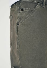 Pantaloni din denim verde măsliniu, cu un croi drept, textură subtilă și un buzunar din spate cu o etichetă de marcă.