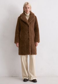 Cappotto lungo marrone e fluffy con colletto a scollatura, chiusura con bottoni e tasche laterali, abbinato a pantaloni a coste crema e scarpe nere.