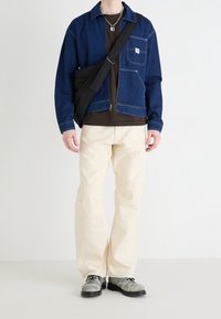 Persona que lleva una chaqueta de mezclilla azul, camisa marrón, pantalones crema, zapatos grises, collar de cadena plateado y bolsa cruzada negra contra un fondo blanco.